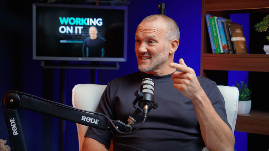 A man sits in a chair with a Rode microphone, recording a podcast in a professional central london studio with a television in the background.